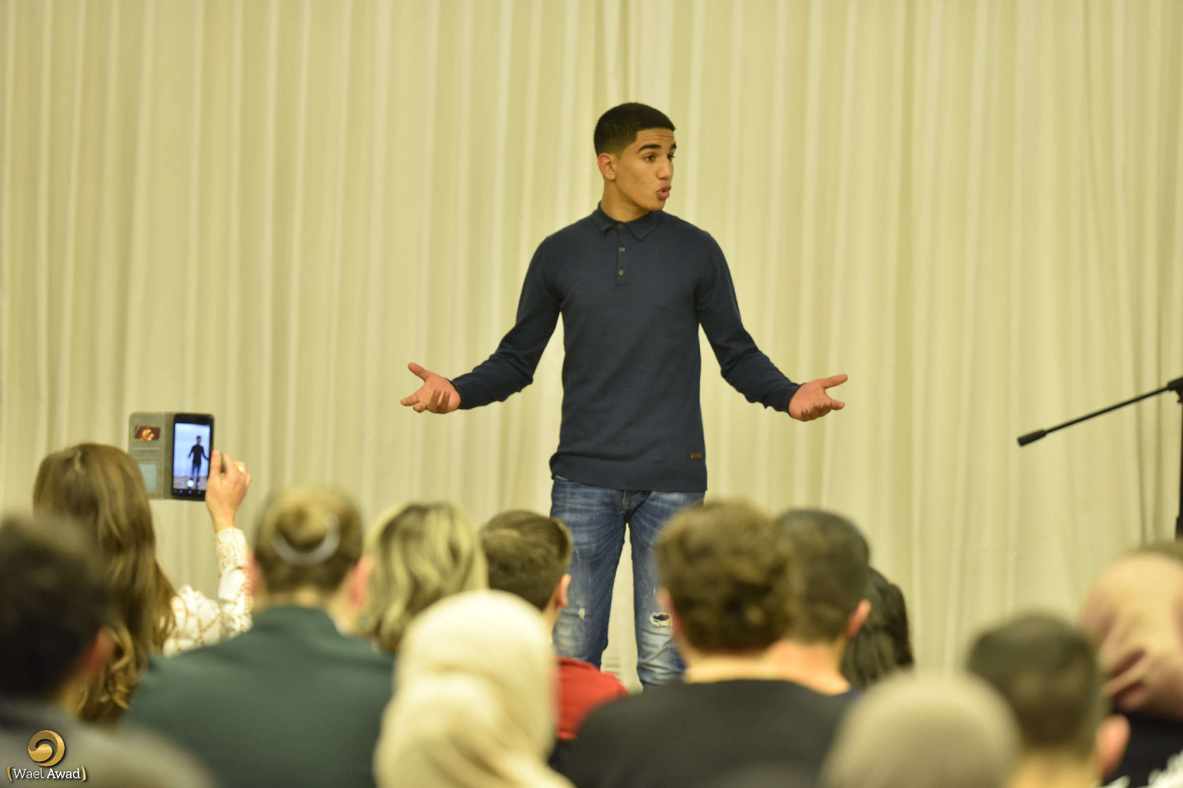 A teenager on stage, arms wide, speaking to an audience.