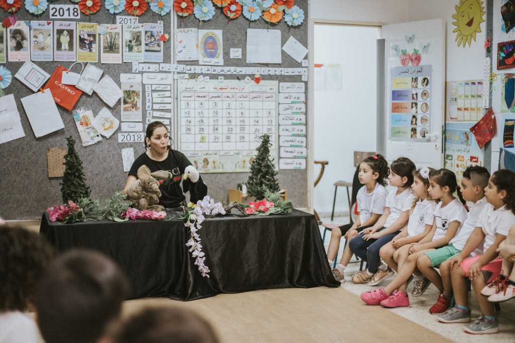A classroom circle: a storyteller with puppets, children engaged, seen from the side.