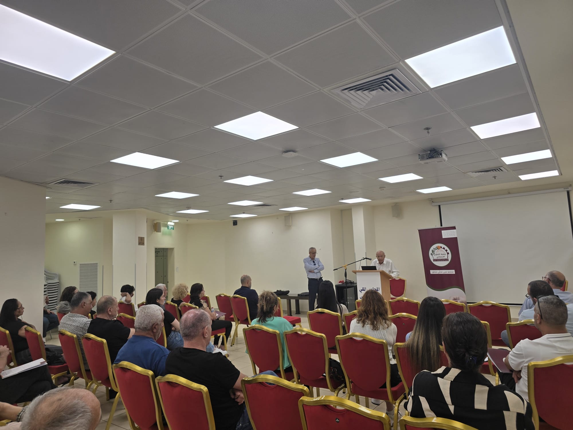 Seated audience in rows facing a speaker at the podium, with the SDC banner behind. Arab Civic Council founding assembly, Haifa, 1 July 2026.